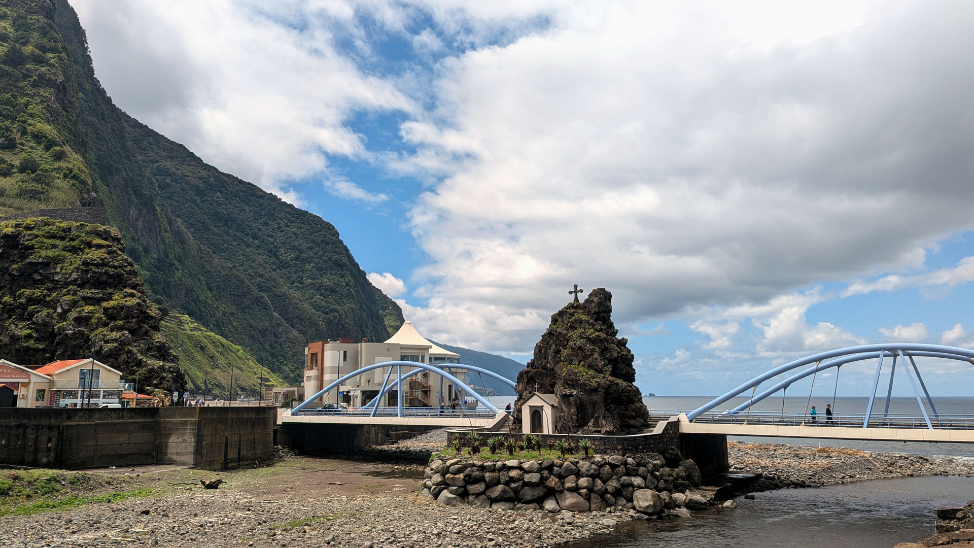 Chapel of São Vicente with the bridge, north‑coast cliffs and calm blue sea under a bright spring sky with cotton clouds