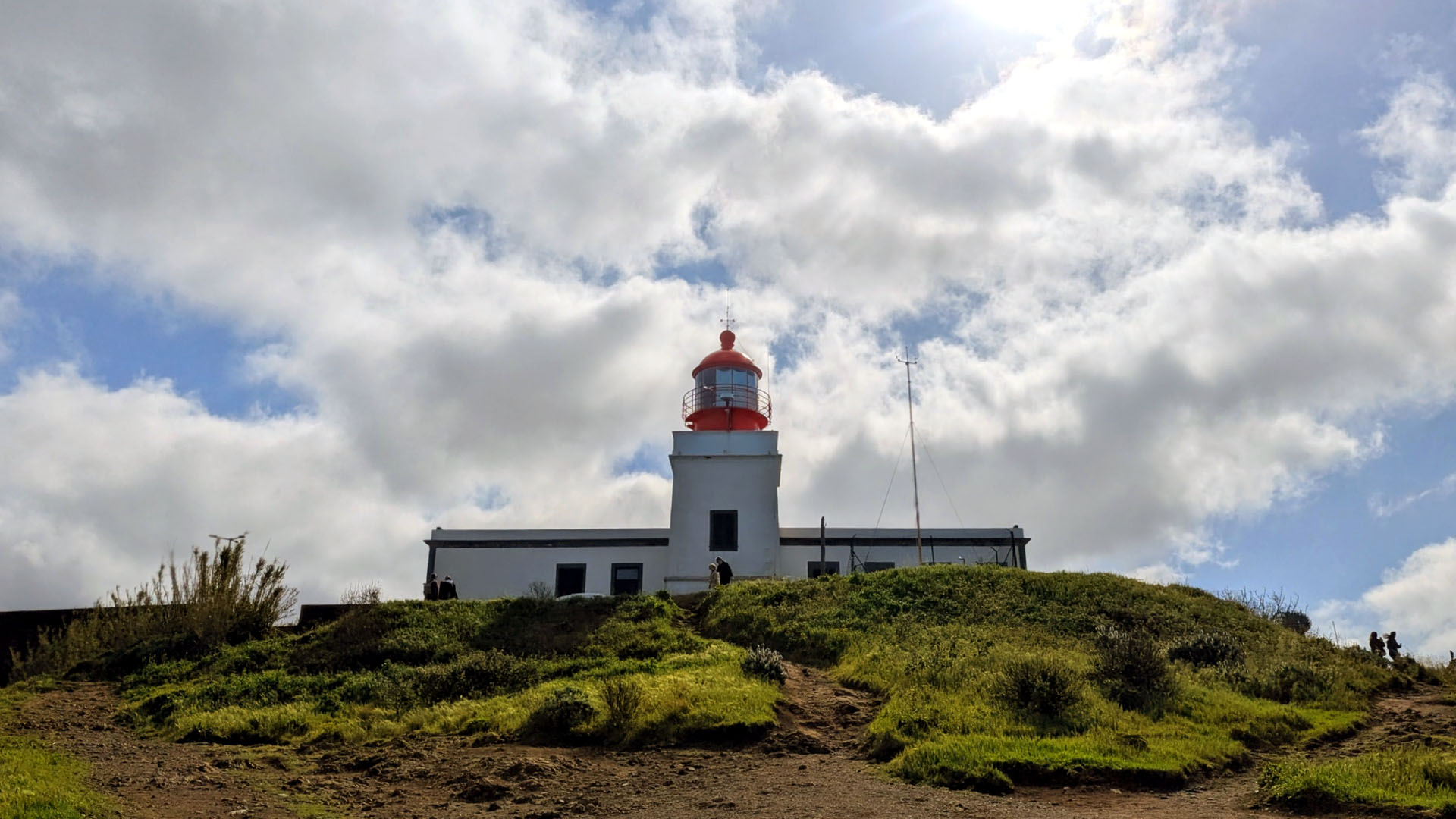 Ponta do Pargo Lighthouse photographed from below, with grass in the foreground, blue sky above and changing spring clouds around the tower